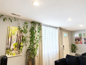 Una sala de estar con un cuadro grande, una planta en maceta, una ventana con cortinas y una puerta.