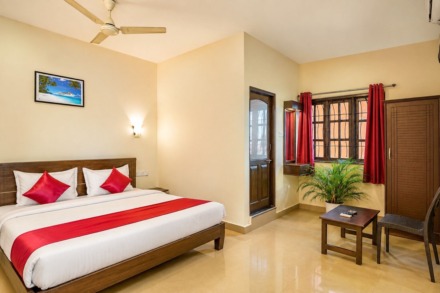 Una habitación de hotel con cama, escritorio, silla, planta en maceta, ventana con cortinas, puerta y ventilador de techo.