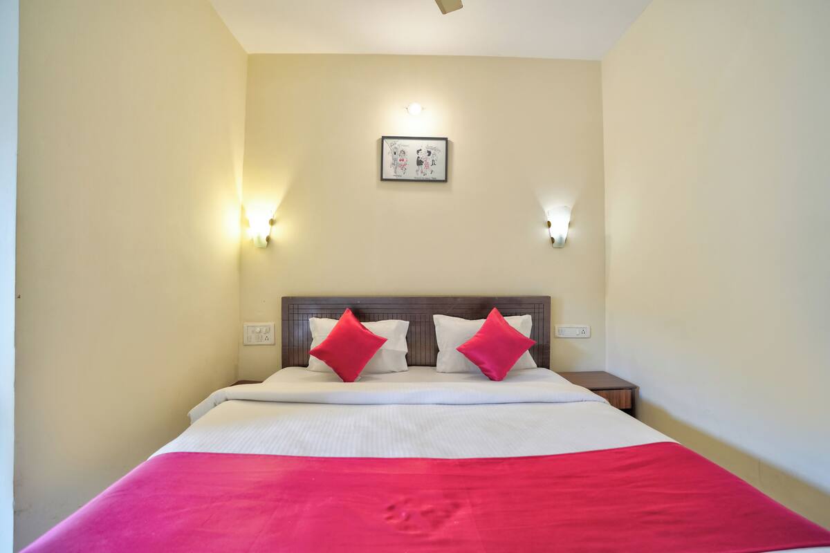 Habitación de hotel con una cama, dos almohadas rojas, una colcha blanca, una manta roja, un cabecero, dos luces fijadas a la pared y un cuadro enmarcado.