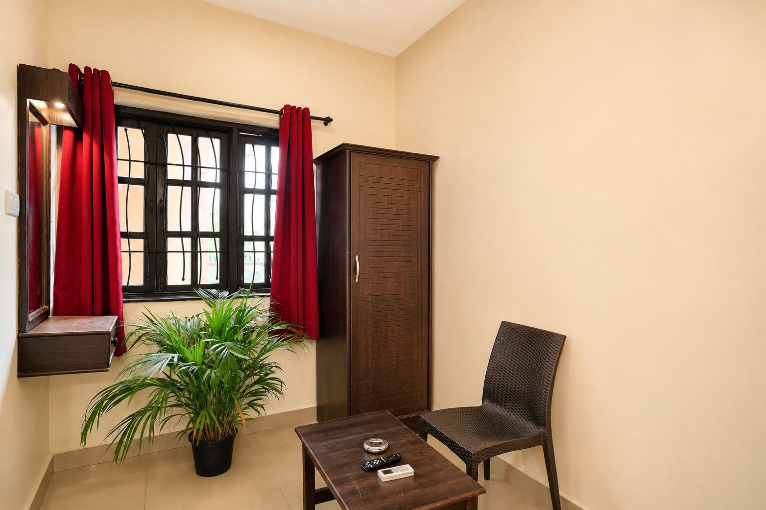 Una habitación pequeña con ventana, una planta en maceta, un armario de madera, una silla de mimbre y una mesita con un control remoto.