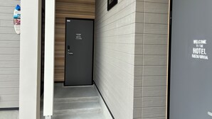Fachada de hotel con diseño moderno, destacando una puerta oscura con un letrero de bienvenida, una escalera y una pared con textura.