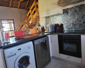 Cocina compacta equipada con lavadora, lavavajillas y horno. Una escalera de madera conduce a un nivel superior.