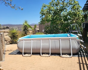 Una piscina inflable con escalera, rodeada por un patio de ladrillo, con vista a edificios lejanos y montañas.