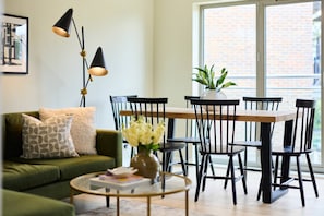Ein Wohnzimmer mit einem grünen Sofa, einem runden Couchtisch, einem Esstisch mit Stühlen und einer Zimmerpflanze.
