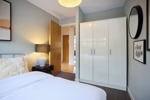 A bedroom with a bed, bedside table, lamp, wardrobe, and a door leading to another room. - Stoke Newington Olive Velvet Three-Bed (Greater London)