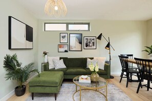 A modern living room with a green sofa, a glass coffee table, and a dining area with black chairs. - Stoke Newington Olive Velvet Three-Bed (Greater London)