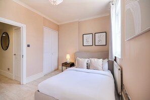 A bedroom with a bed, a nightstand with a lamp, a door, and two framed pictures on the wall. - Olive Velvet & Marble - South Kensington (Greater London)