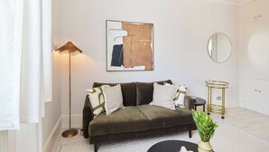 En stue med en sofa, en gulvlampe, et rundt spejl, et lille bord med en plante, og et indrammet abstrakt billede.
