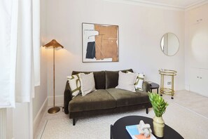 A living room with a sofa, a floor lamp, a round mirror, a small table with a plant, and a framed abstract painting. - Olive Velvet & Marble - South Kensington (Greater London)