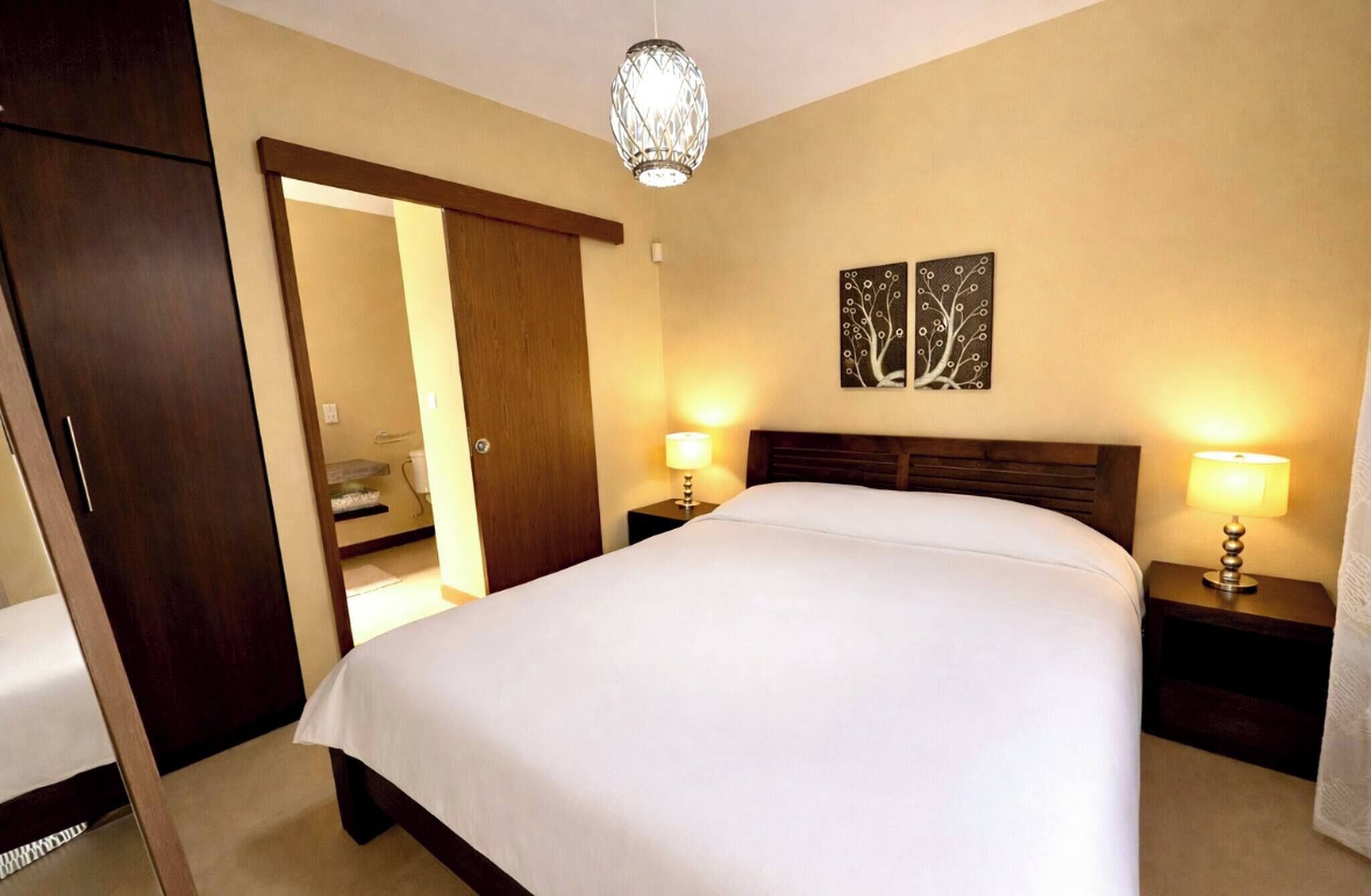 A hotel room with a bed, two bedside lamps, a wooden headboard, a door, and a wall-mounted artwork.