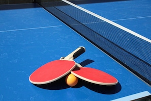 A blue ping pong table with two red paddles and a yellow ball.