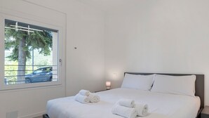 Une chambre moderne avec un grand lit, deux tables de chevet, une fenêtre donnant sur une voiture et des arbres, et une petite table de chevet avec une lampe.