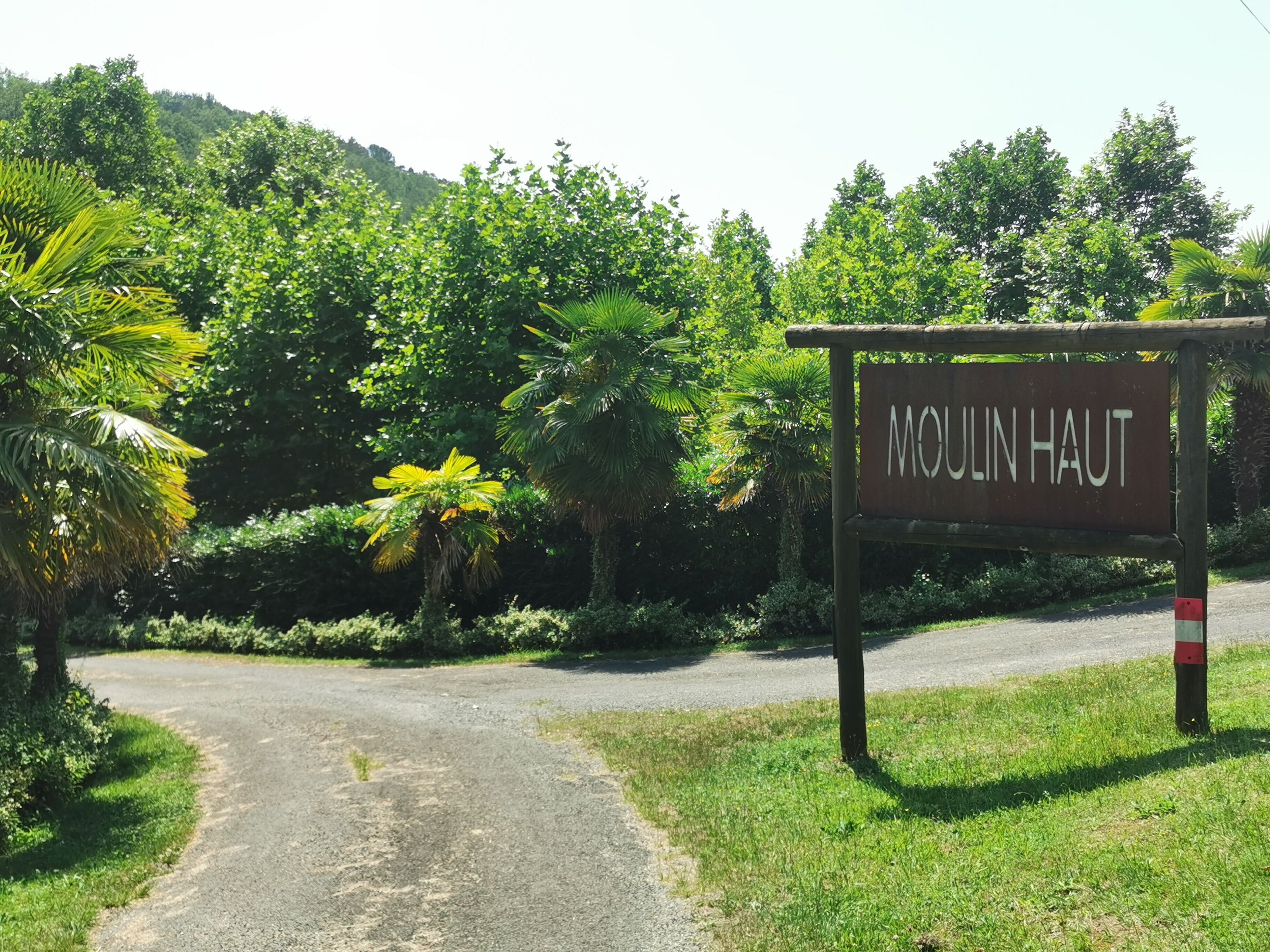 Ein Schild mit der Aufschrift 'Moulin Haut' am Eingang einer von üppigem Grün umgebenen Straße.