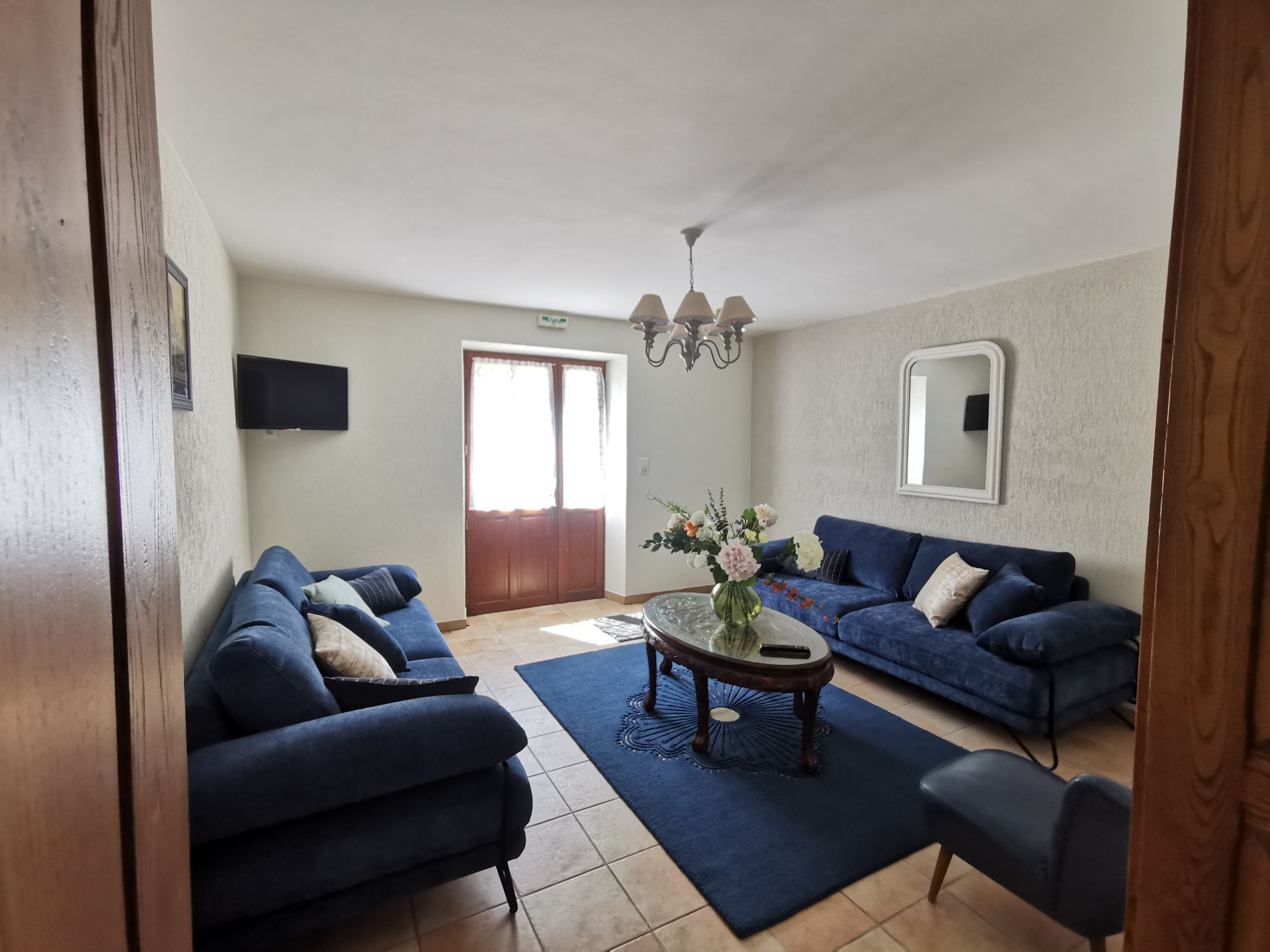 Un salon avec deux canapés bleus, une table basse sur laquelle est posé un vase, un téléviseur fixé au mur et une porte donnant sur l’extérieur.