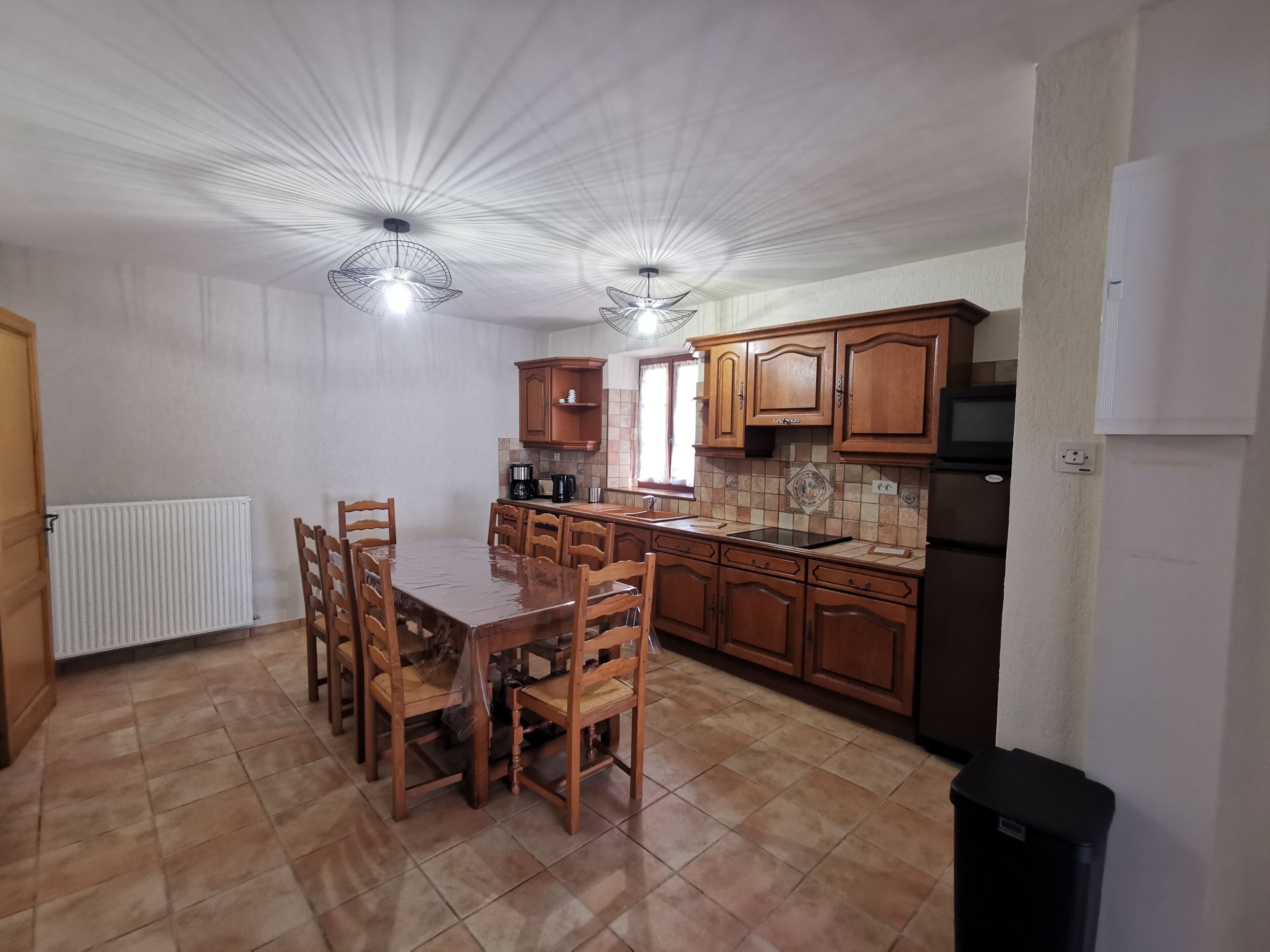 A kitchen with wooden cabinets, a dining table, chairs, a refrigerator, and a black trash bin.