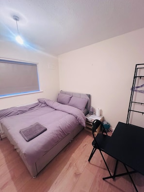 A bedroom with a bed, a desk, a chair, a radiator, a window with blinds, and a ceiling fan.