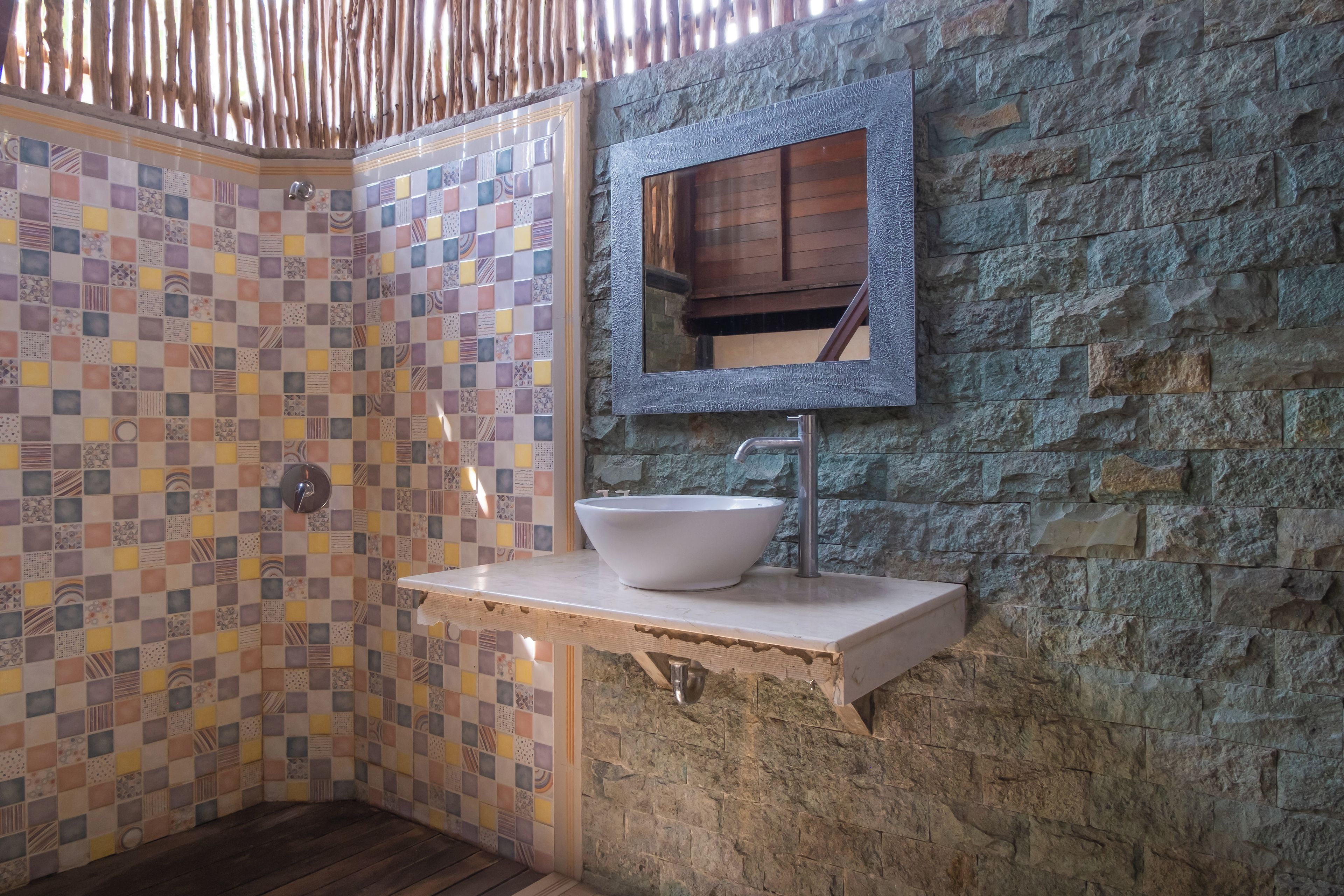 A bathroom with a tiled shower wall, a stone wall, a sink with a mirror, and a wooden floor.