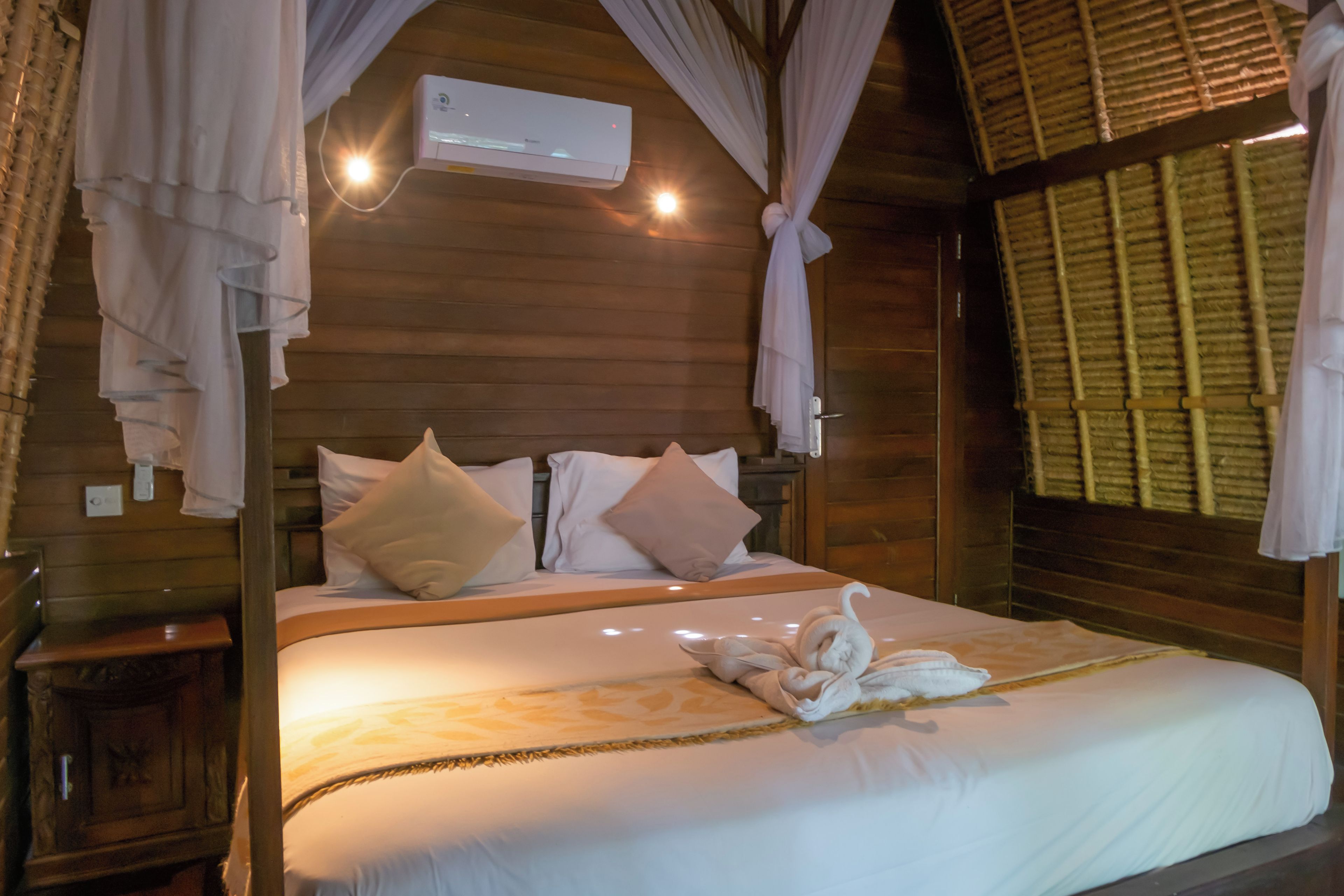 Una habitación de madera con una cama, ropa de cama blanca, un aire acondicionado de pared y una ventana con persiana de bambú.