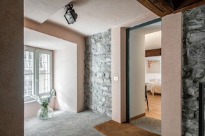 Ein Zimmer mit einer Steinwand, einer Holzbalkendecke, einem Fenster mit Jalousien und einer Tür, die in einen anderen Raum führt.