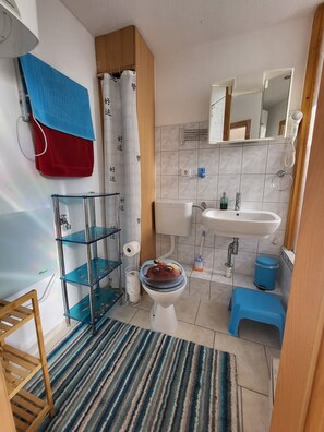 A bathroom with a toilet, sink, shower curtain, and a blue trash bin. - Vacation apartment Zwergenstübchen (Lübstorf)