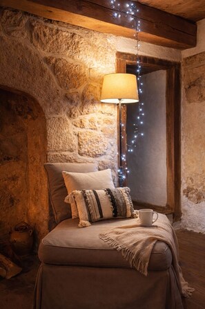 Una habitación acogedora con pared de piedra, techo de madera, una lámpara, un cojín y una taza sobre una mesa.