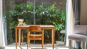 Uma mesa de madeira com uma cadeira em frente a uma janela de vidro, parede de bambu e um sofá cinzento.