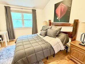 A bedroom with a bed, a desk, a dresser, a window with curtains, and a painting on the wall.