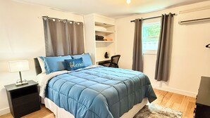 Quarto com cama, mesa de cabeceira, escrivaninha e janela com cortinas.
