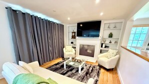 Uma sala de estar com sofá, poltronas, mesa de centro, televisão de tela plana, lareira e estante de livros.