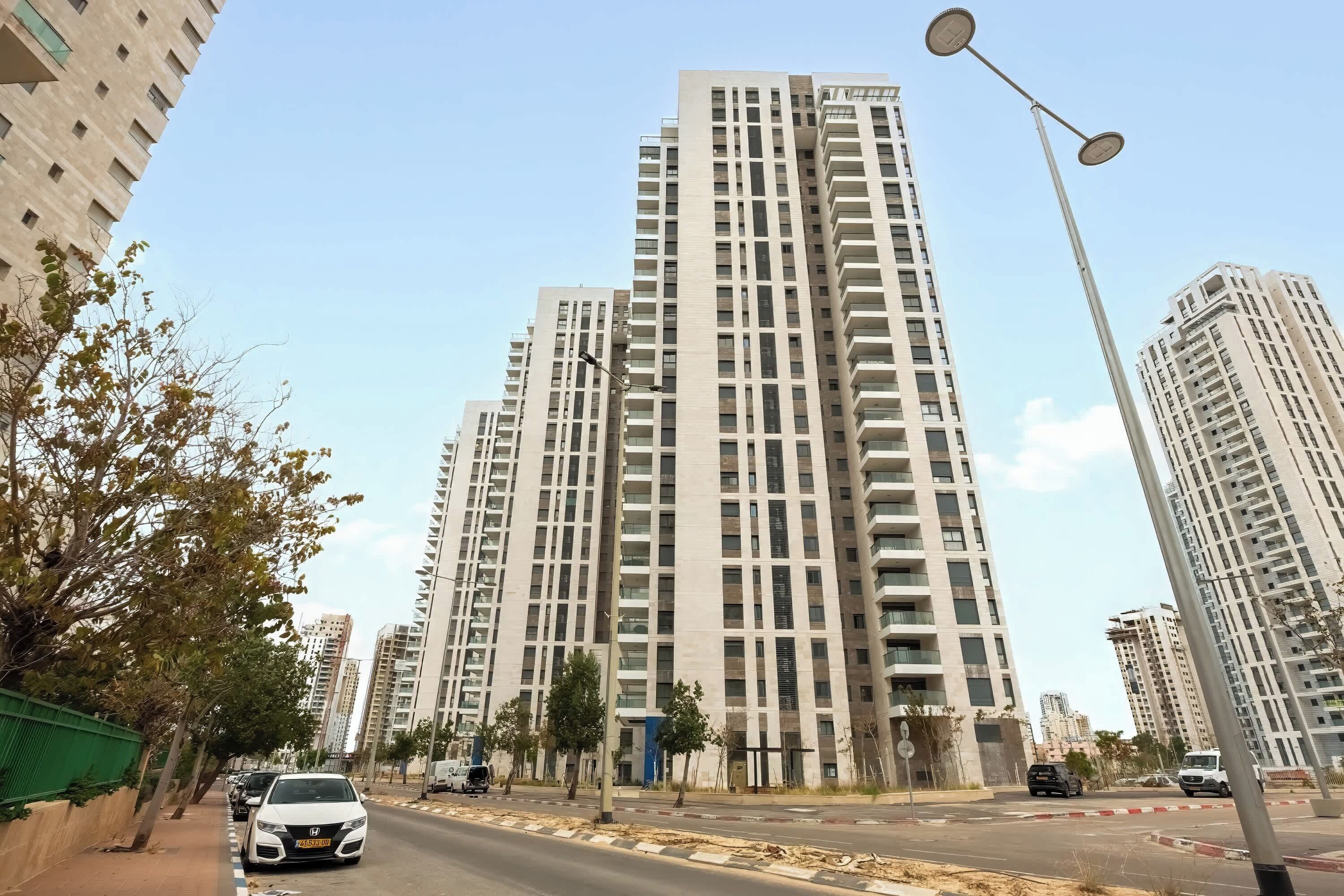 Un edificio residencial alto con varios pisos y balcones, cielo despejado y una calle con coches estacionados.