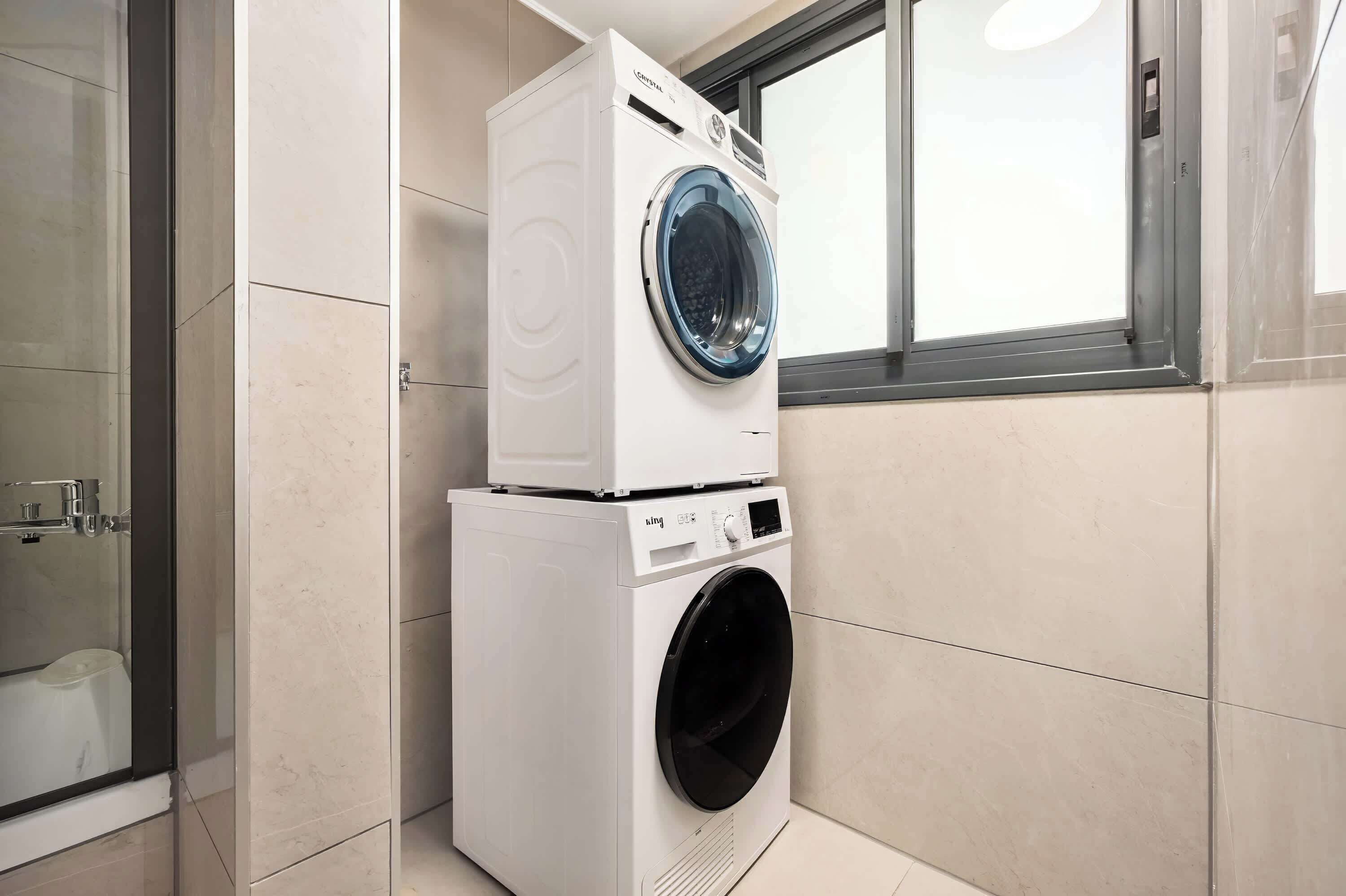 A compact laundry area with a front-loading washer and dryer stacked on top of each other, a glass shower enclosure, and a window providing natural light.