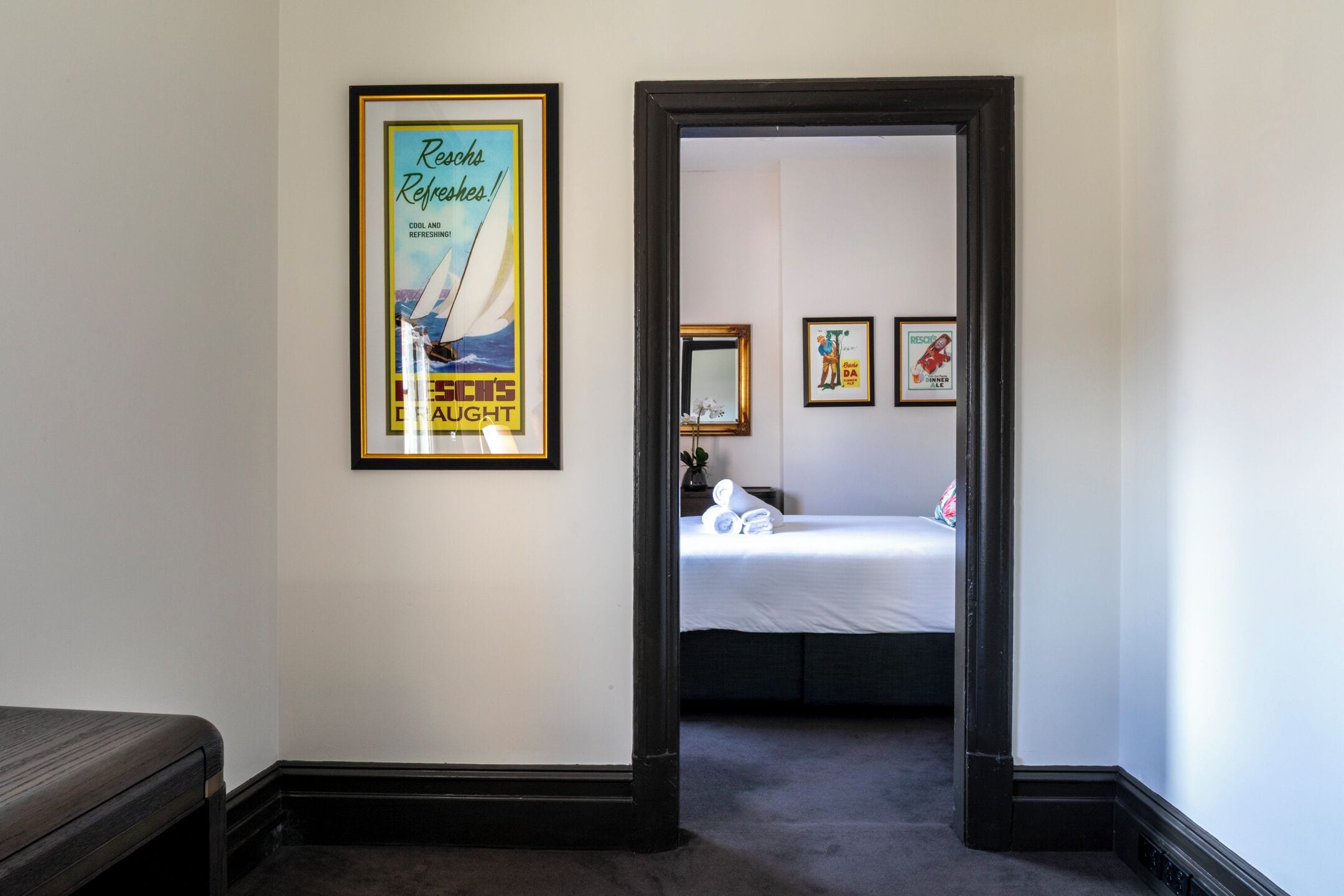 A hotel room with a bed, a framed poster on the wall, and a doorway leading to another room.