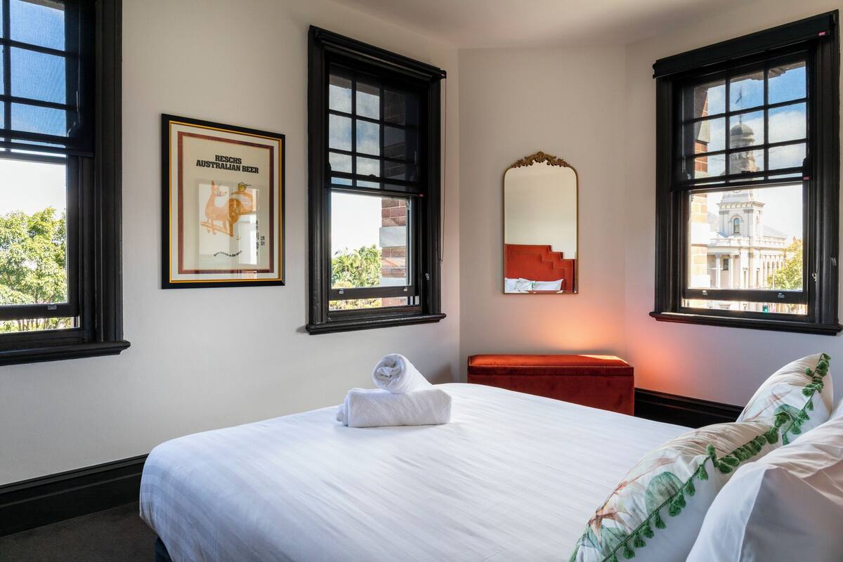 A hotel room with a bed, a red bench, a framed picture, and windows with a view of a building.