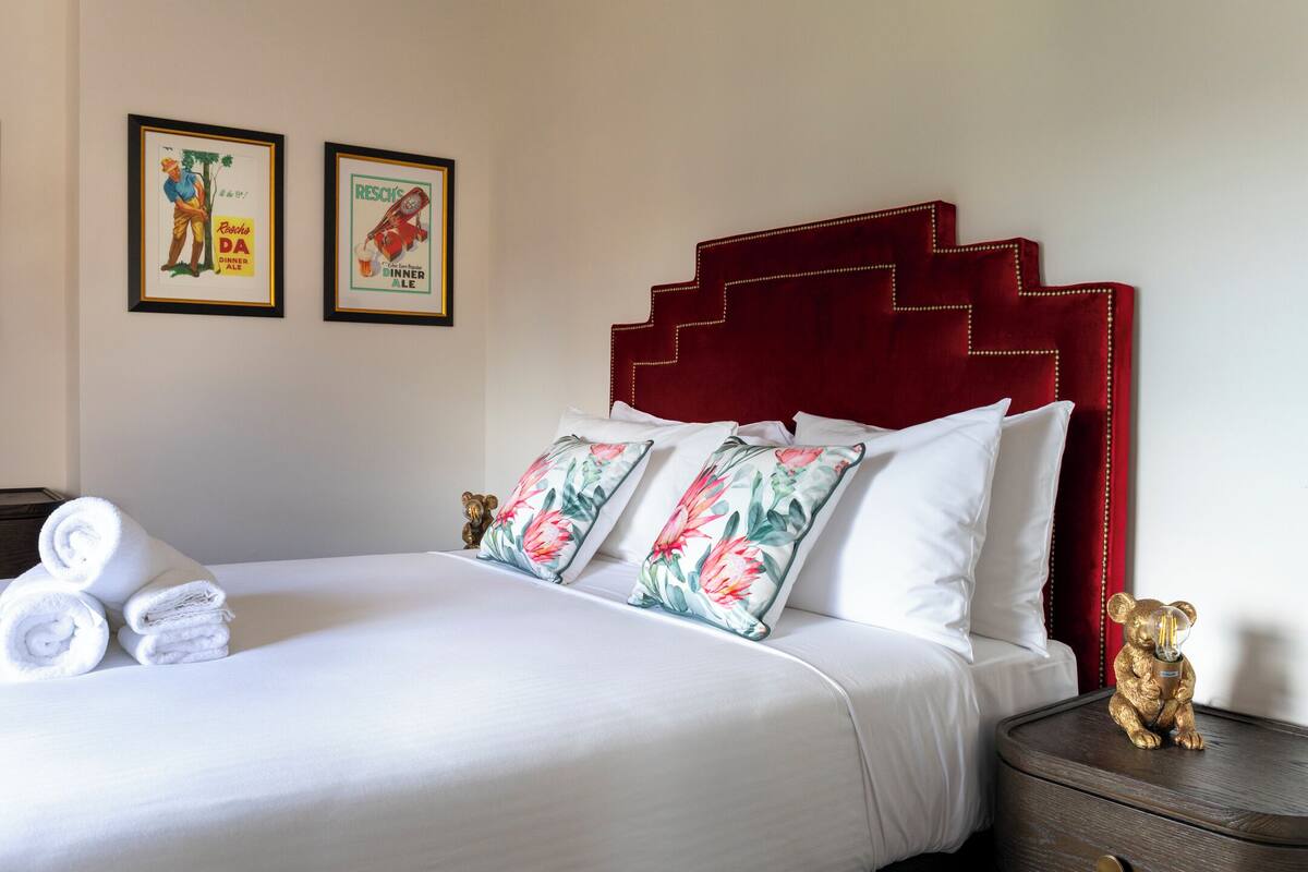 A neatly made bed with white linens and floral pillows, a red upholstered headboard, and two framed pictures on the wall.