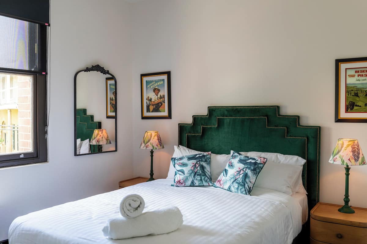 A neatly made bed with a rolled white towel, a green upholstered headboard, and a framed poster on the wall.