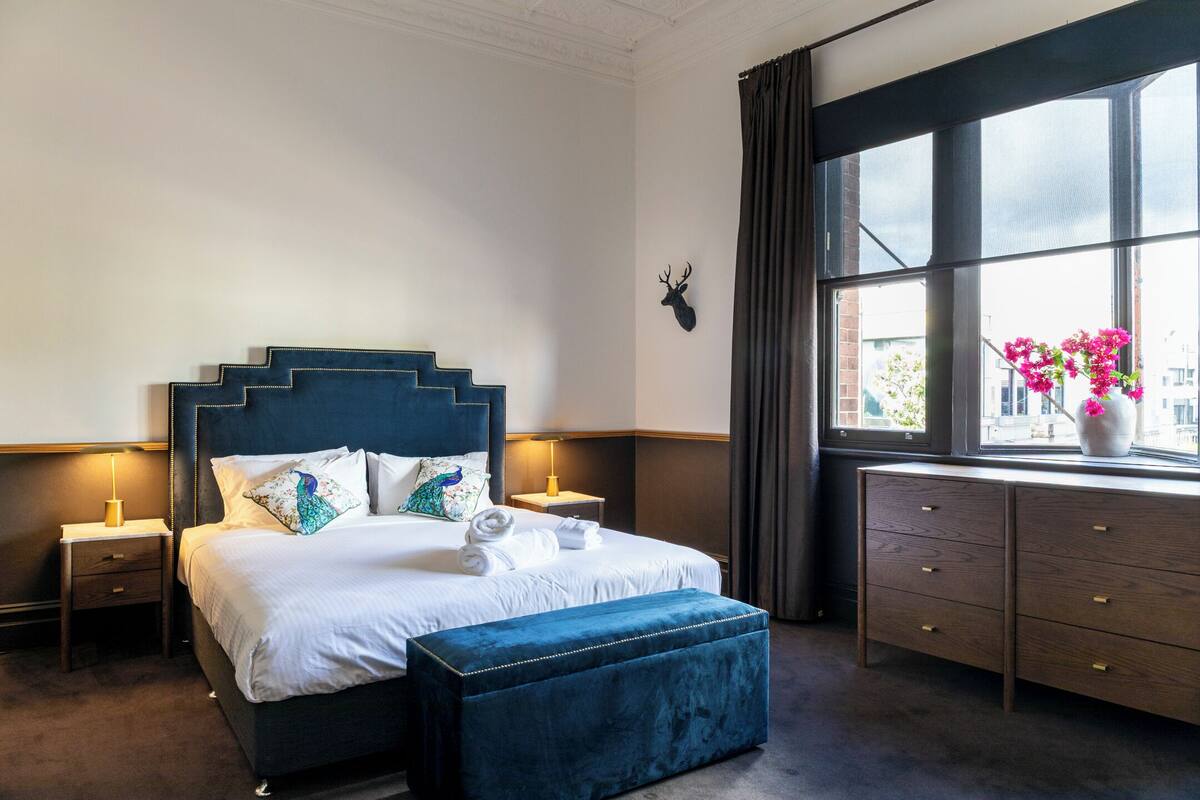 A hotel room with a bed, bedside tables, a dresser, a window with curtains, and a wall-mounted deer head decoration.