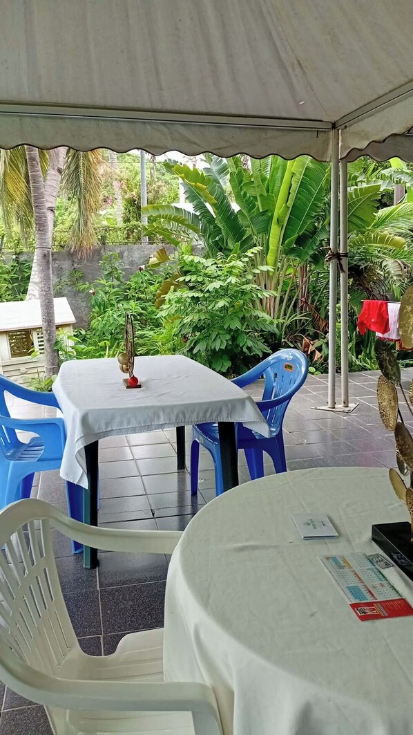An outdoor dining area with tables and chairs under a canopy, surrounded by lush greenery.