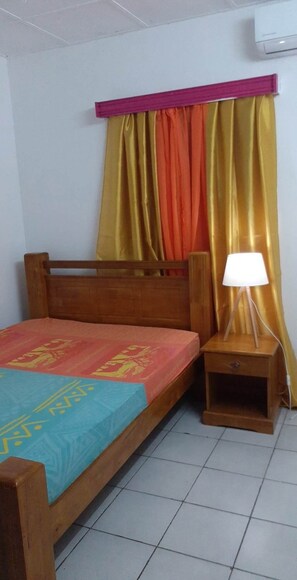 A single bed with a wooden frame, a bedside table with a lamp, and curtains in a room with white walls and a tiled floor.