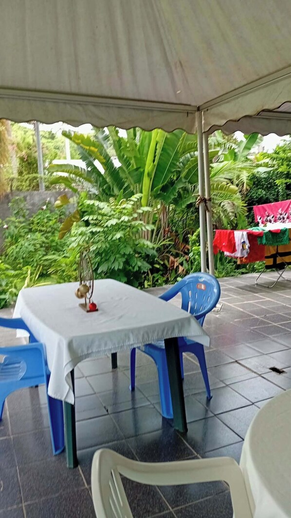 Eine Terrasse im Freien mit Tisch und StĂŒhlen unter einem Zelt, umgeben von GrĂŒnpflanzen.