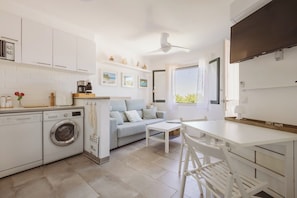A compact kitchen with a washing machine, a dining area with a table and chairs, a sofa, and a ceiling fan. - Apartment with pool dofins 6 (Ciutadella)