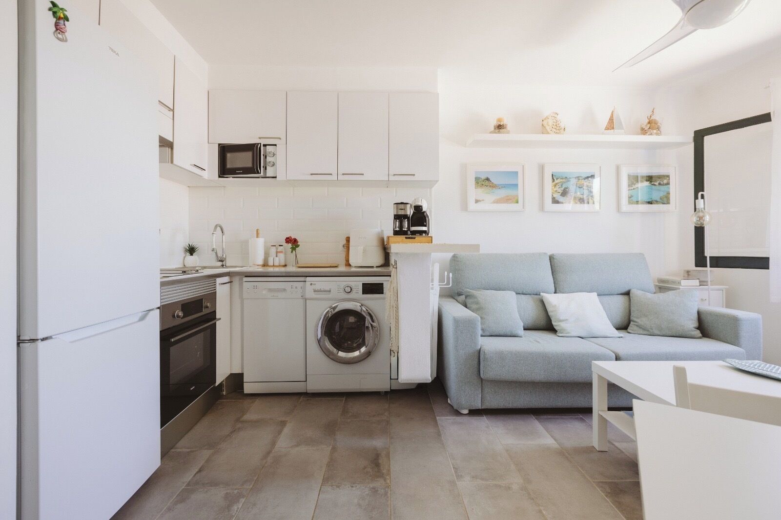 A compact living space with a kitchenette, a washing machine, a grey sofa, and a dining area.