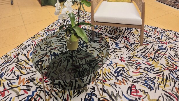 Ein Zimmer mit einem Sessel, einem Tisch mit gemustertem Tischlein, einer Topfpflanze, einem Vorhang und einer farbenfrohen Glasscheibe.