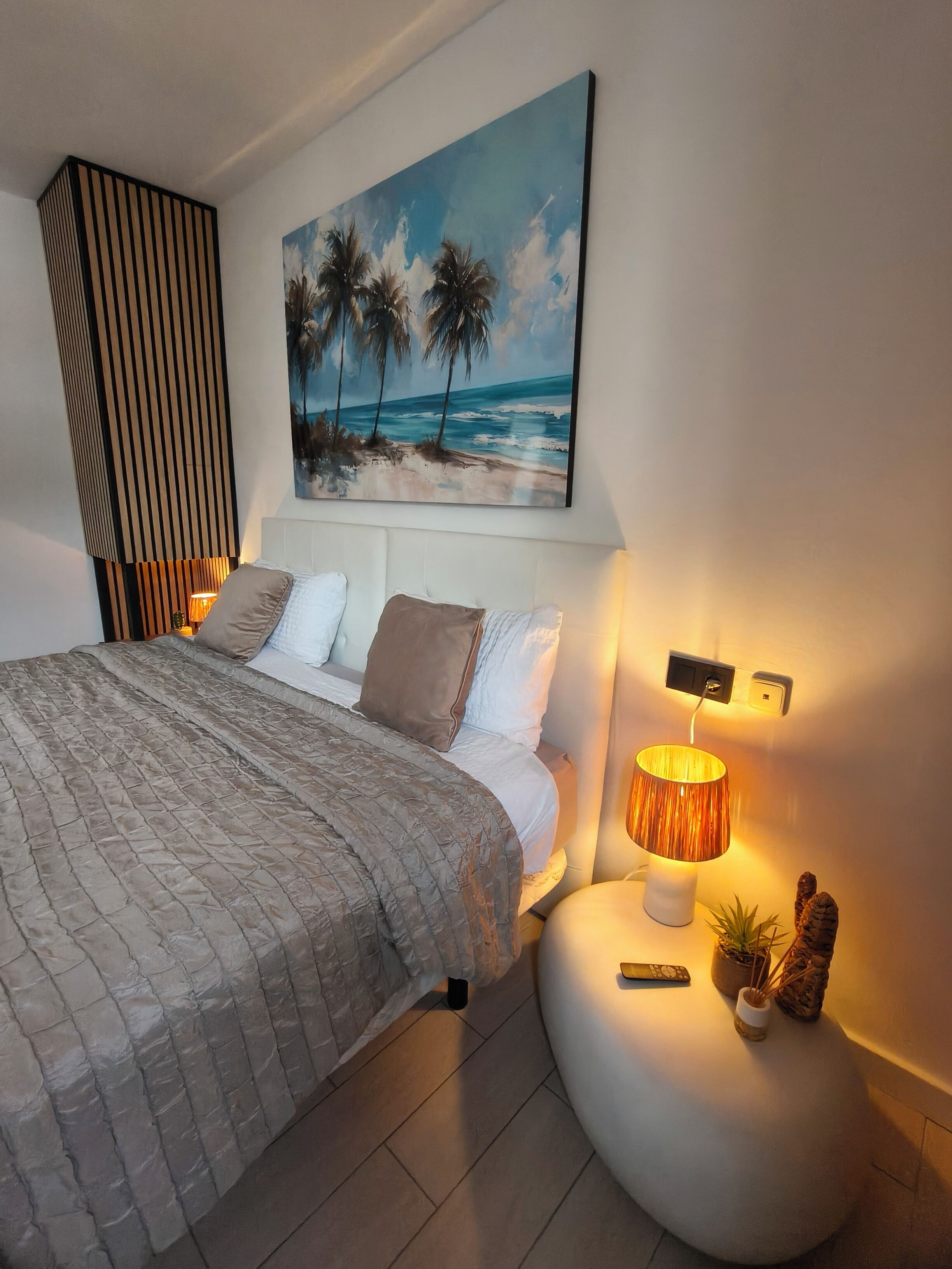 A bedroom with a bed, a painting of a beach, a bedside lamp, and a small plant on a nightstand.
