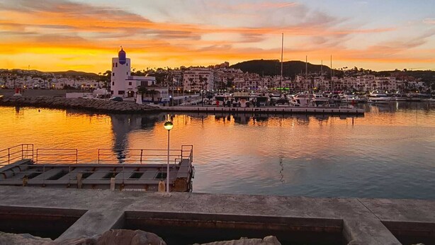 Una cittadina costiera al tramonto con un porto turistico, edifici e una torre bianca imponente.
