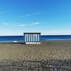 Ein Strand mit einer kleinen, blau und weiß gestrichenen Struktur, vermutlich ein Rettungsschwimmerstandort, vor einem Sandstrand mit dem Meer im Hintergrund.