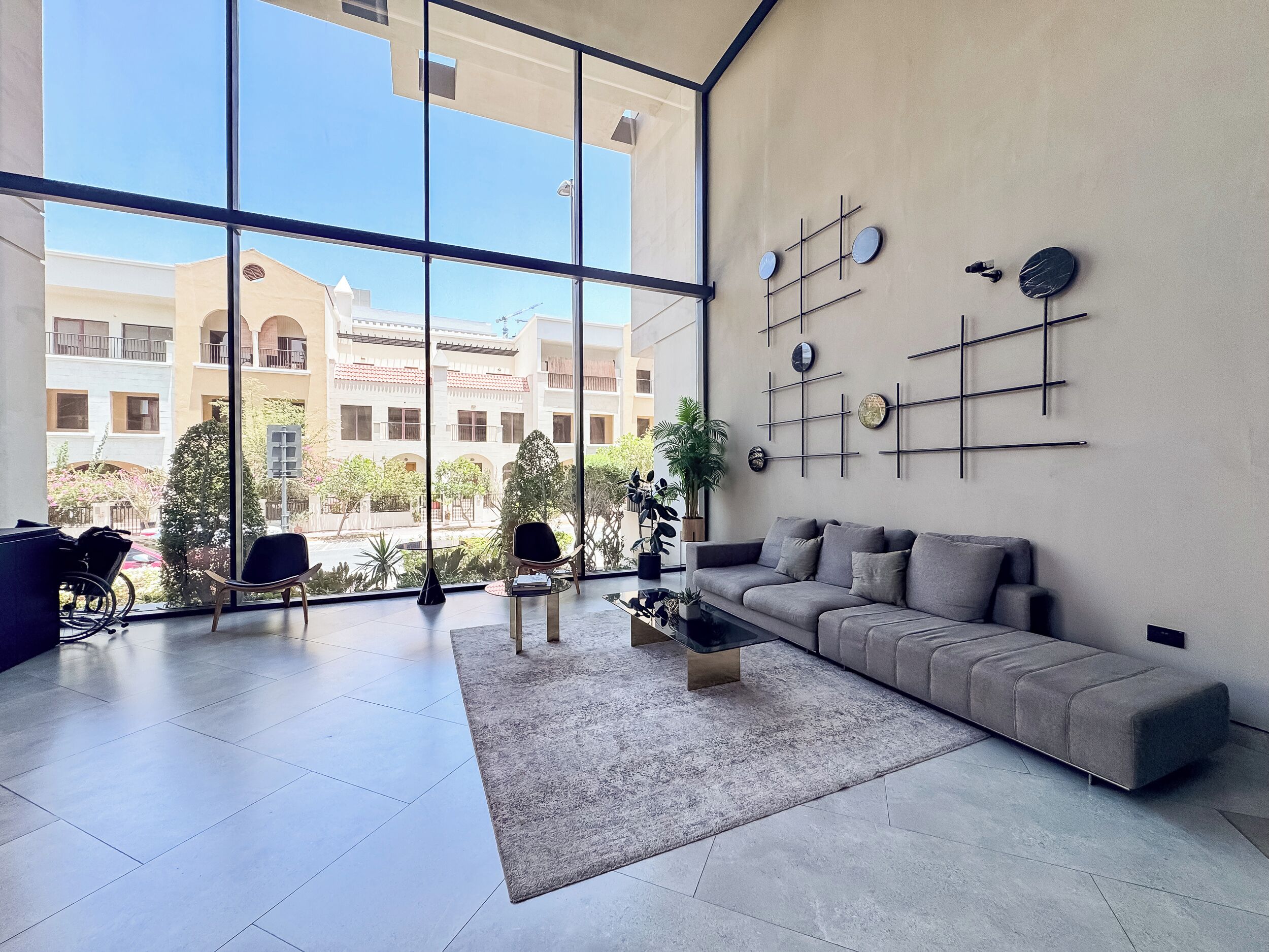 A modern living room with large windows, a grey sofa, a coffee table, and a wall-mounted clock.
