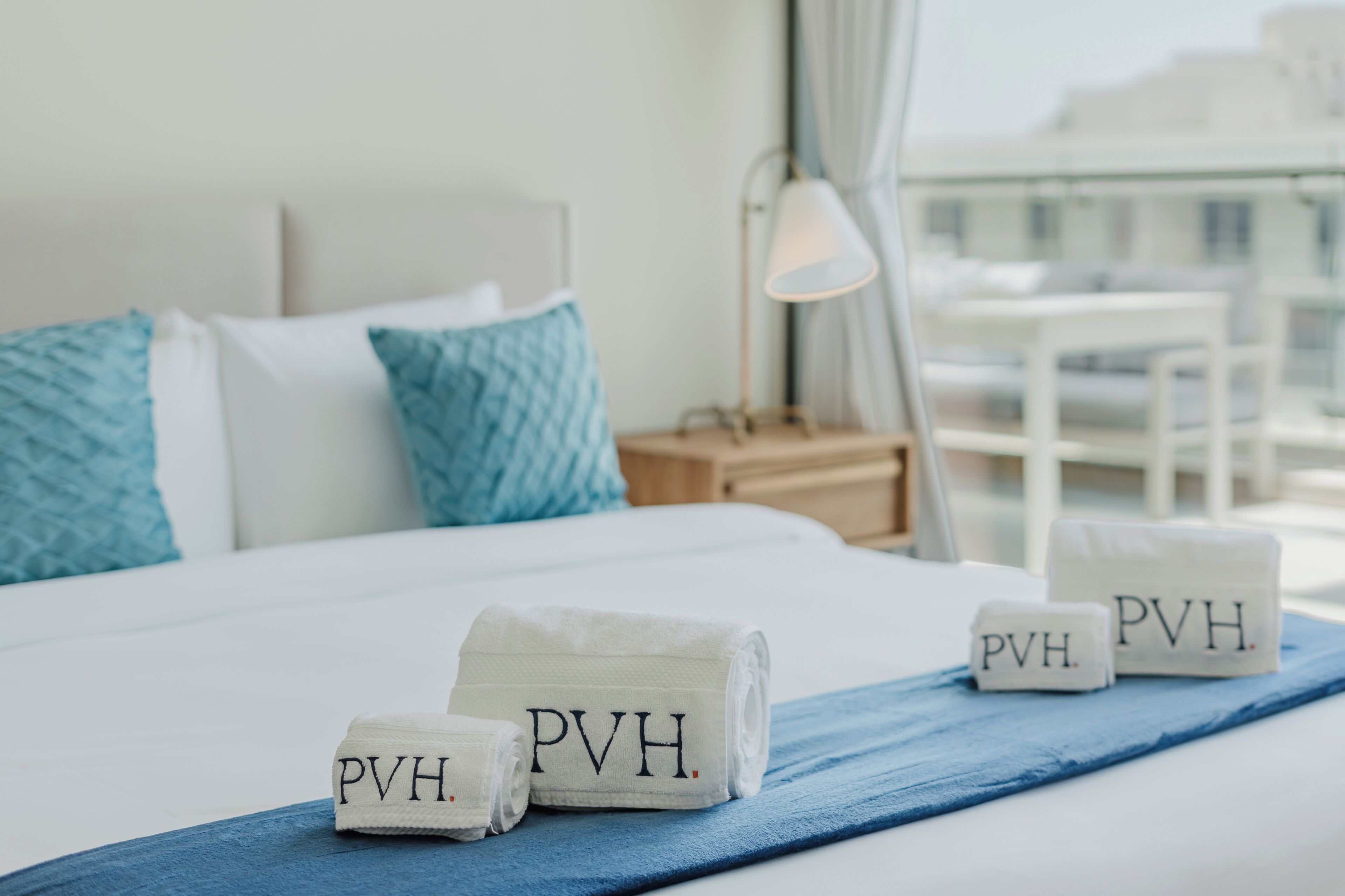 Un lit bien fait, recouvert d’une courtepointe bleue, avec des oreillers et deux serviettes brodées du sigle « PVH ».