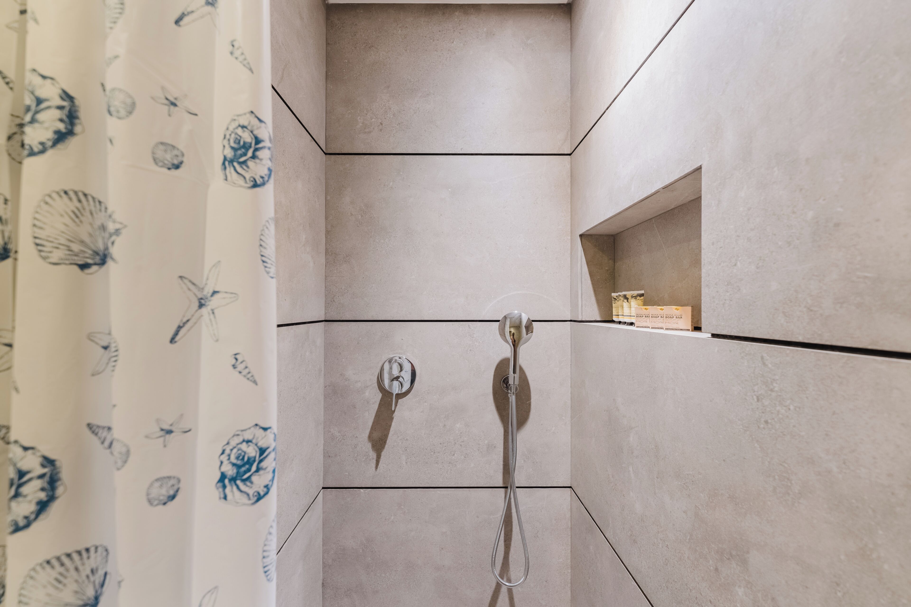 A shower with a handheld showerhead, a wall-mounted control, and a shelf with a box.