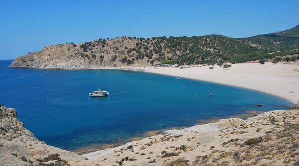 Un paysage côtier avec un bateau sur l’eau, une plage de sable et une colline rocheuse à la végétation clairsemée.