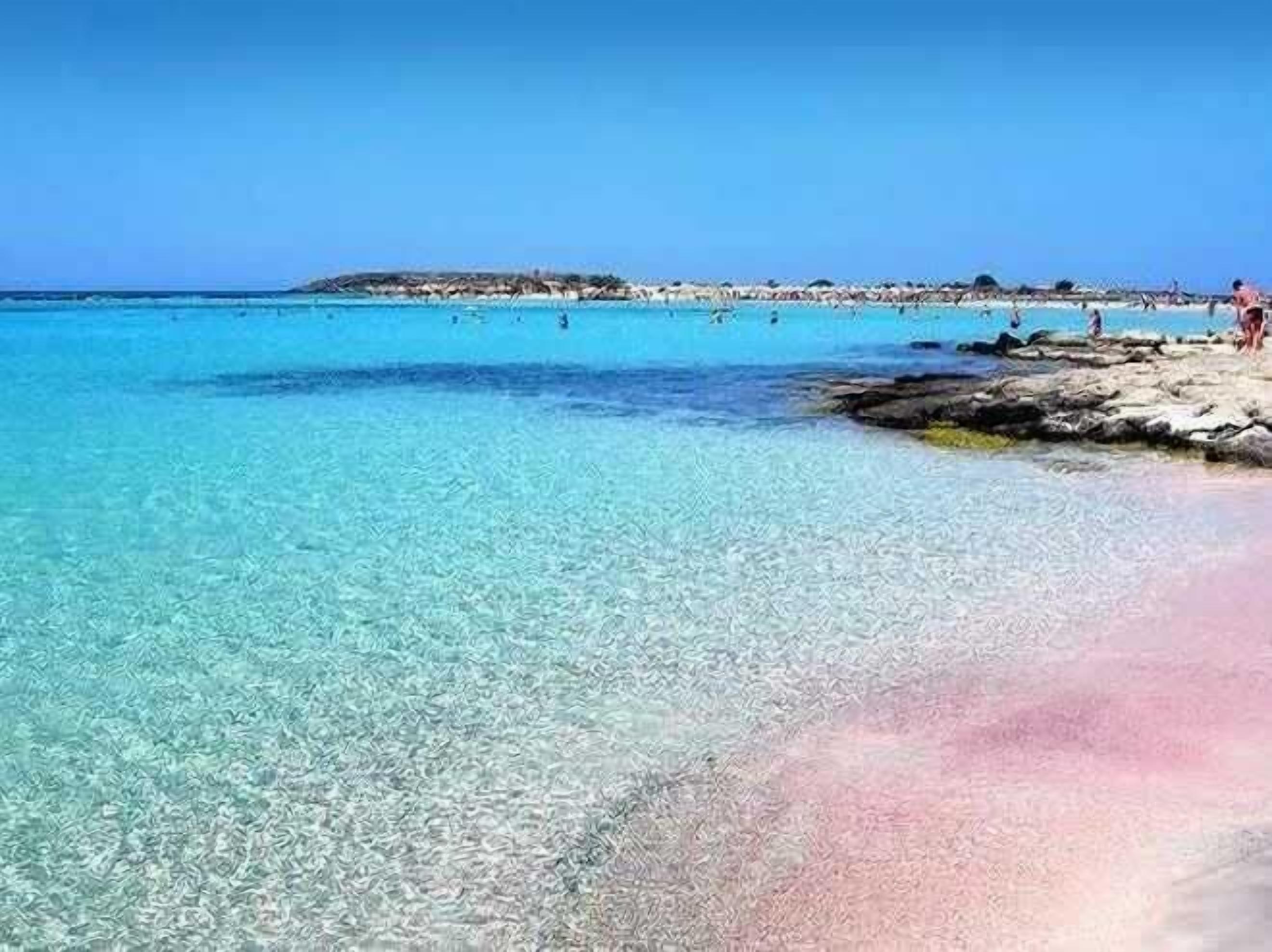 Une plage bordée d’une eau turquoise et limpide, d’une côte rocheuse et d’une terre visible au loin.